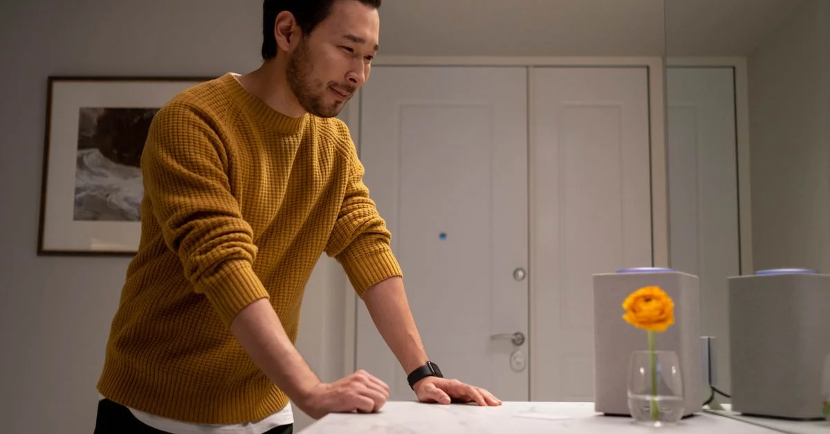 Un homme en pull moutarde souriant à une enceinte intelligente dans un cadre intérieur moderne.