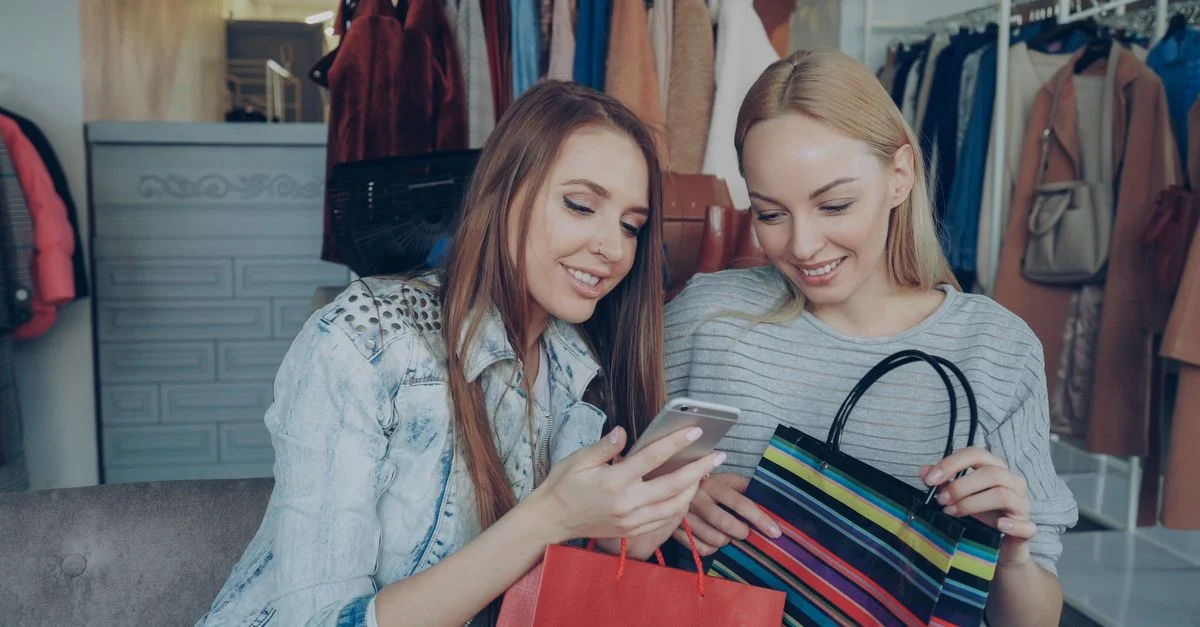 Deux femmes profitant d'une séance de shopping et partageant un moment sur leur smartphone dans un magasin de vêtements.